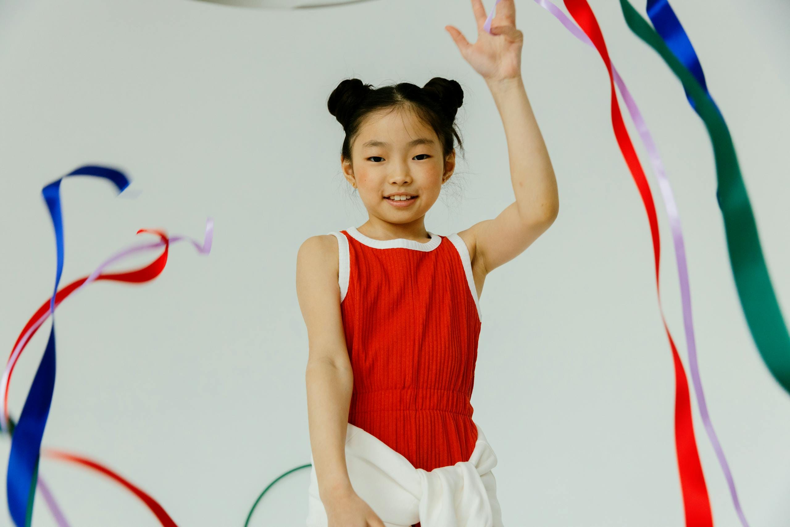 Cheerful young girl in a red dress joyfully dancing with colorful ribbons on a white background.