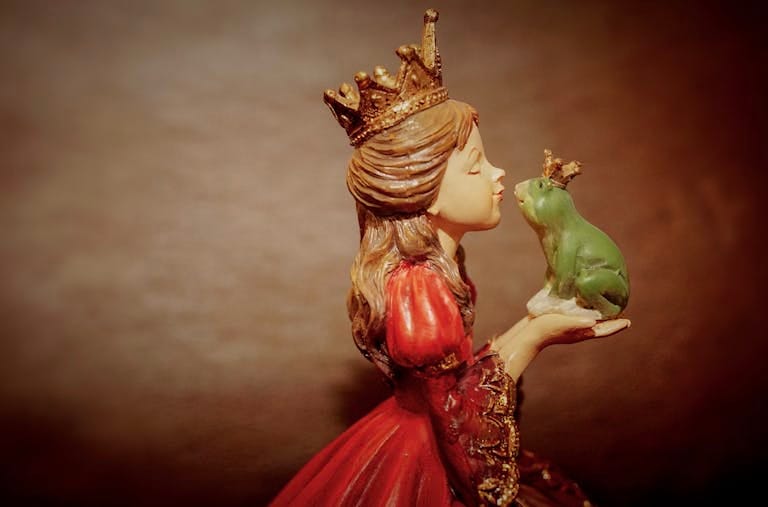 Side view of a ceramic princess figurine holding and kissing a crowned frog, evoking fairy tale magic.